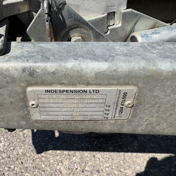 Close-up of a dusty metal suspension component with a maker's plate and bolts in view, showing worn gray metal and a small amount of legible text on the plate.