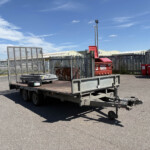 Industrial yard scene with a flatbed trailer carrying metal fencing panels and a red dumpster; blue sky above and warehouse buildings in the background