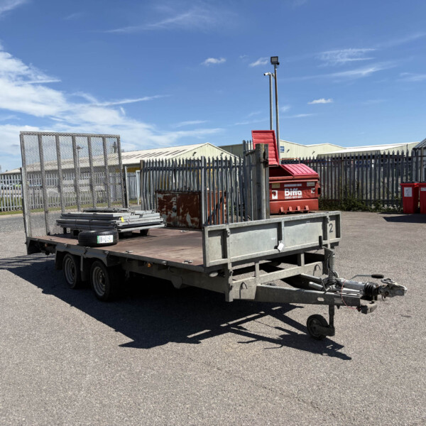 Industrial yard scene with a flatbed trailer carrying metal fencing panels and a red dumpster; blue sky above and warehouse buildings in the background
