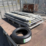 Trailer carrying stacked metal frames and a spare tire, with a chain-link fence and rusted cabinet in a storage yard in the background.
