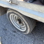 Close-up of a metal trailer wheel and tire on asphalt, dusty with dirt and grime.