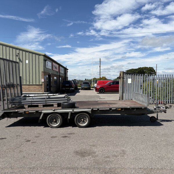 Flatbed trailer in a parking lot beside a green metal-clad warehouse, with blue sky above and a chain-link fence on the left.