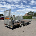 Trailer loaded with metal fencing panels in a parking lot, red vans in the background against a blue sky with clouds.