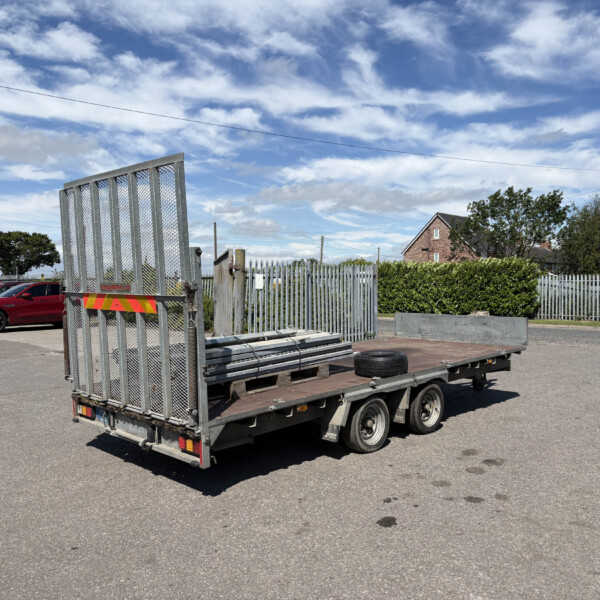 Trailer loaded with metal fencing panels in a parking lot, red vans in the background against a blue sky with clouds.