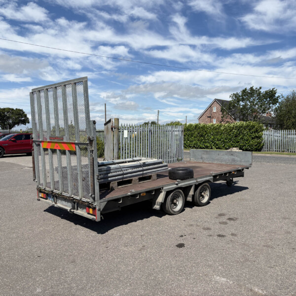 Silver metal flatbed trailer with a tall ramp in a parking lot; red cars in the background and a blue sky with clouds behind.
