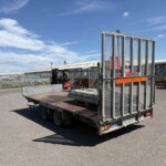 Industrial yard with a flatbed trailer and tall metal mesh gate, forklift in the background under a blue sky.