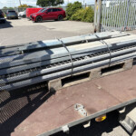 Flatbed trailer carrying stacked galvanized metal beams strapped down in a parking lot near a fence. In the background are parked cars and a red vehicle.