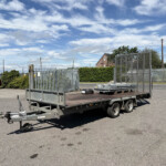Flatbed trailer with metal mesh sides parked in an empty lot, blue sky and scattered clouds overhead, and a red car in the background.