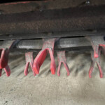 Close-up of red curved metal tines from agricultural equipment hanging over a dusty concrete floor, with a dark rusted beam above.