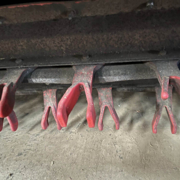 Close-up of red curved metal tines from agricultural equipment hanging over a dusty concrete floor, with a dark rusted beam above.