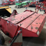 Red farm machinery in a storage yard with other equipment visible in the background, including a yellow attachment on the left and a green machine in the distance