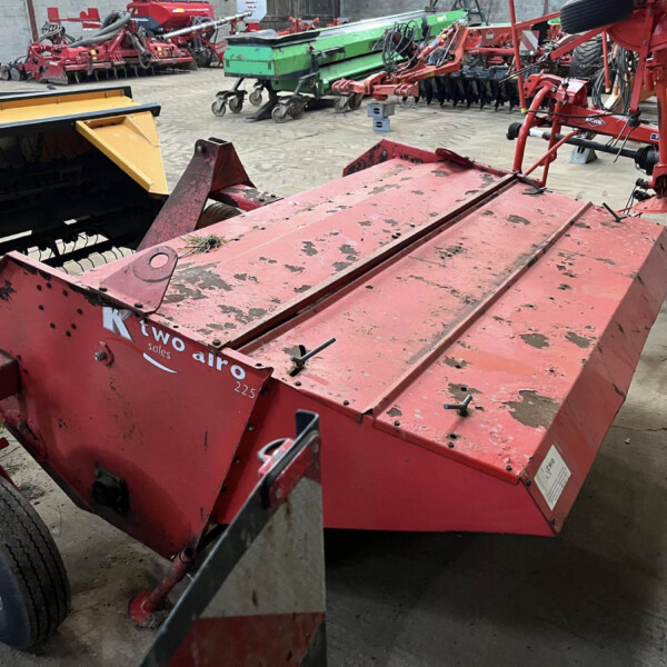Red farm machinery in a storage yard with other equipment visible in the background, including a yellow attachment on the left and a green machine in the distance