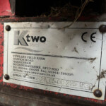 Metal nameplate on a dirty red surface showing 'ktwo sales' and contact details, weathered and stained with soil and cobwebs.