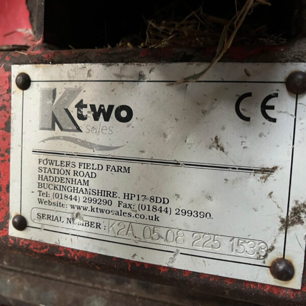 Metal nameplate on a dirty red surface showing 'ktwo sales' and contact details, weathered and stained with soil and cobwebs.
