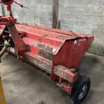 Red farm implement (baler) with rotating tines, parked against a concrete block wall; shows mud, grass debris, and worn paint with a visible wheel.