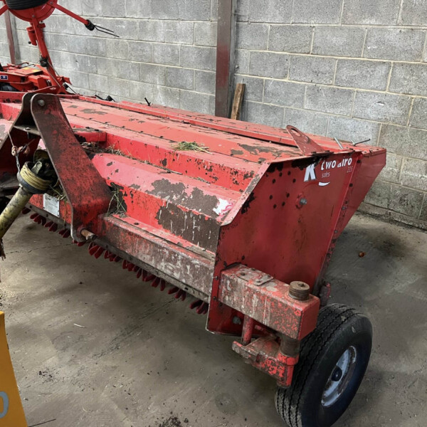Red farm implement (baler) with rotating tines, parked against a concrete block wall; shows mud, grass debris, and worn paint with a visible wheel.