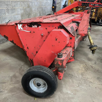 Red, worn-looking agricultural header on a wheeled frame inside a workshop with a gray cinder block wall behind it.