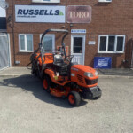 Orange Kubota ride-on lawn mower parked in front of a brick building with Russell's and Toro signs and a reception door nearby.