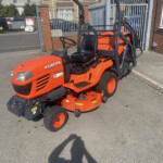 Orange Kubota riding mower parked on a driveway in front of a metal gate and brick house behind it.