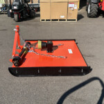 Red industrial mower deck viewed from above in a paved lot, with pallets, machinery, and a large cardboard crate in the background.