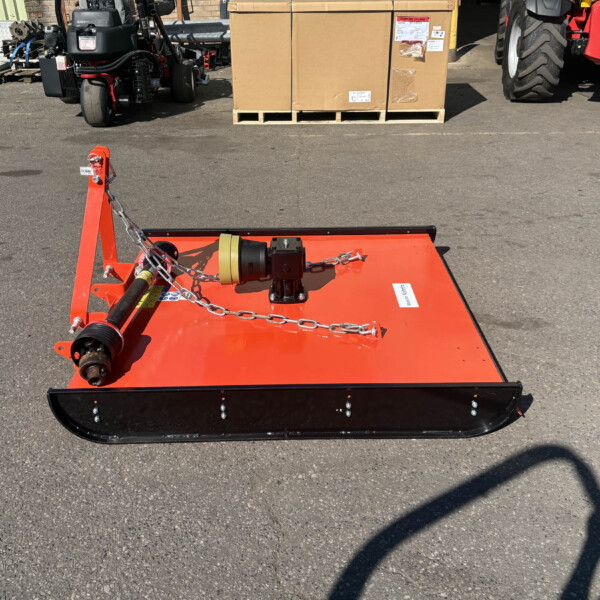 Red industrial mower deck viewed from above in a paved lot, with pallets, machinery, and a large cardboard crate in the background.