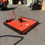Close-up of a red/orange mower deck with chains and hitch on a paved outdoor yard surface.