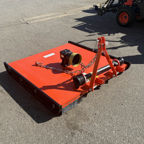 Orange tractor-mounted mower attachment on a paved surface, with chains and a hitch, ready for use; machinery in the background.