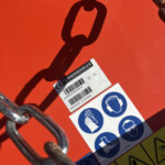 Close-up of an orange industrial surface with a white safety label and a rusted metal chain attached nearby.