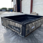 Camouflage-patterned metal trailer in a concrete yard near a warehouse, with a green tractor partially visible on the left.