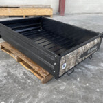 Black metal cargo bed resting on wooden pallets in a concrete, industrial space, no people visible.