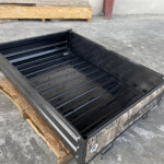 Open black metal toolbox/tray with a ribbed floor on a concrete surface, adjacent to wooden pallets and a concrete wall.