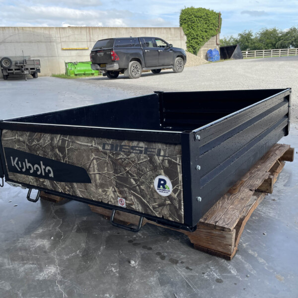 Black camouflage Kubota utility trailer in the foreground, placed on wooden pallets in an industrial lot, with a dark pickup truck and concrete wall in the background.