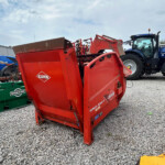 Red Kuhn agricultural implement in the foreground on a gravel lot, with a blue tractor in the background.
