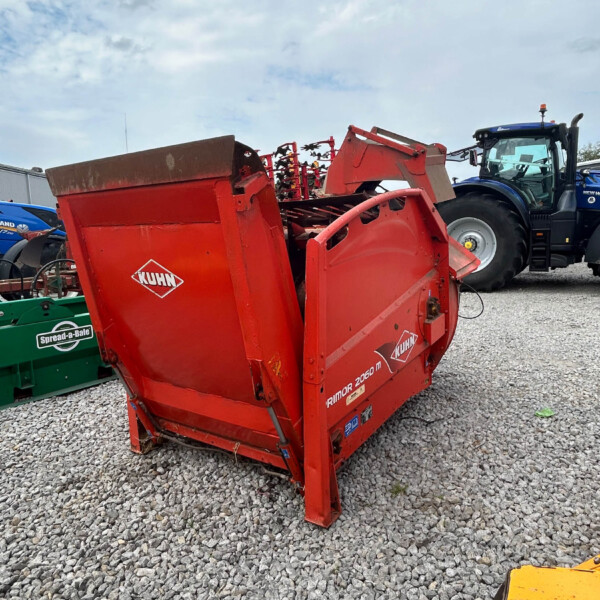 Red Kuhn agricultural implement in the foreground on a gravel lot, with a blue tractor in the background.