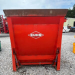 Red KUHN agricultural hopper/spreader in an outdoor equipment lot on a gravel surface, blue sky above