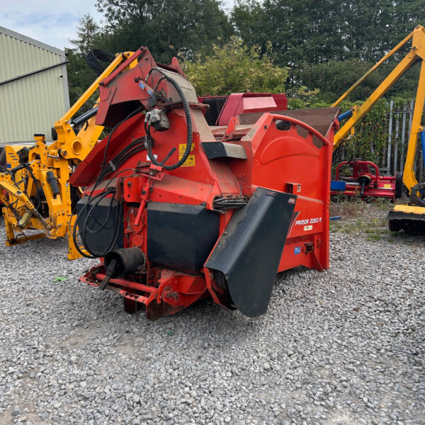 Red agricultural baler with yellow hydraulic arms parked on a gravel lot, outdoors near trees and a fence, hoses visible