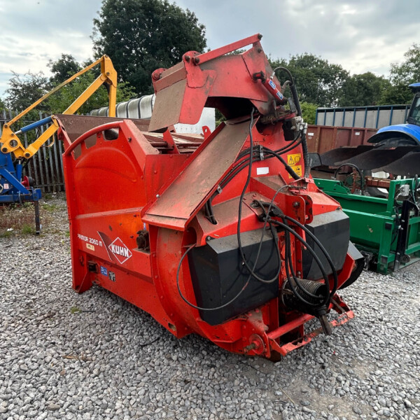 Red agricultural machine with hoses and a chute in a gravel lot, backed by other machinery and trees in the distance