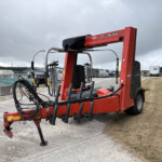 Red Kuhn SW 4014 bale wrapper trailer with hydraulic arms and hoses in an outdoor yard under cloudy sky