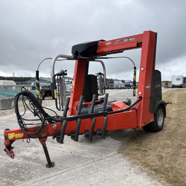 Red Kuhn SW 4014 bale wrapper trailer with hydraulic arms and hoses in an outdoor yard under cloudy sky