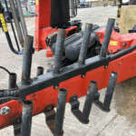 Close-up of a red construction machine with several black, curved tines or forks and hydraulic hoses at a work site, dirt ground in the background