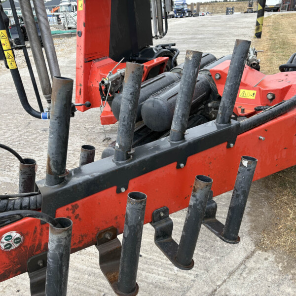 Close-up of a red construction machine with several black, curved tines or forks and hydraulic hoses at a work site, dirt ground in the background