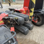 Red agricultural/industrial machine with large black tires and yellow-black warning stripes; close-up of hydraulic components and hoses on a dirt surface