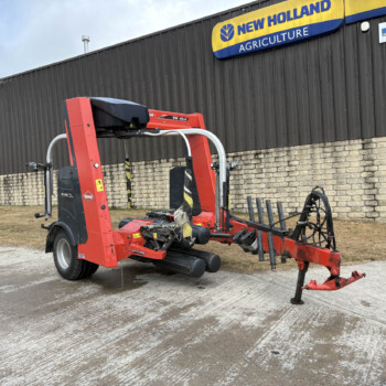 Red agricultural baler/trailer equipment parked in a lot beside a dark metal building with a New Holland Agriculture sign above it.