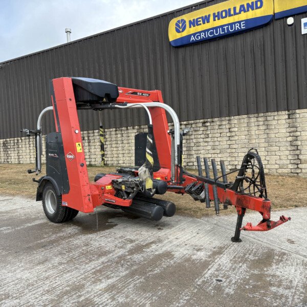 Red agricultural baler/trailer equipment parked in a lot beside a dark metal building with a New Holland Agriculture sign above it.