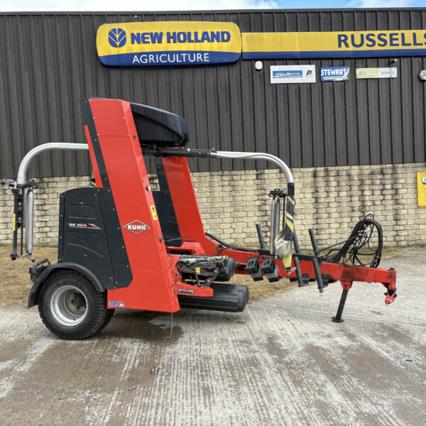 Red KUHN agricultural baler parked on a concrete yard in front of a New Holland Agriculture building sign.