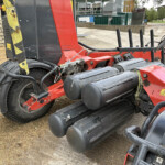 Red industrial machine with black rollers and hoses outdoors, close to a stone wall and grassy area.
