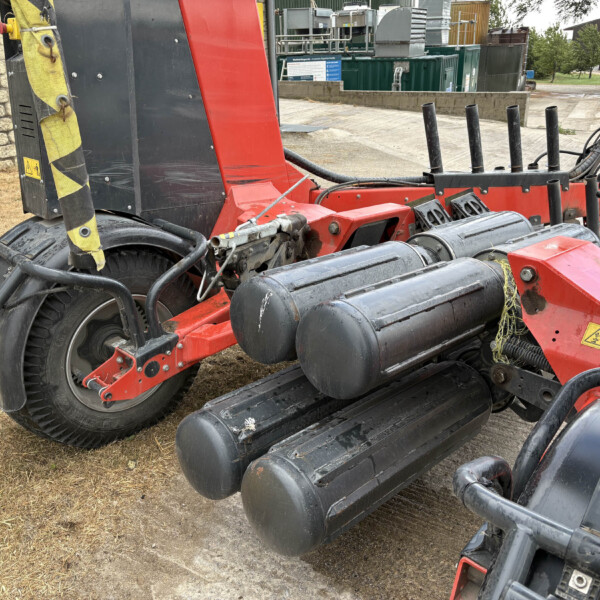 Red industrial machine with black rollers and hoses outdoors, close to a stone wall and grassy area.