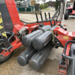 Red agricultural machine with several black vertical rollers and hoses, parked beside a tractor outdoors in a yard.