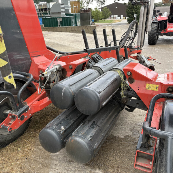 Red agricultural machine with several black vertical rollers and hoses, parked beside a tractor outdoors in a yard.