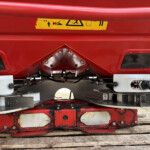 Red industrial machine underside with a yellow safety label and metal brackets visible on a wooden platform.